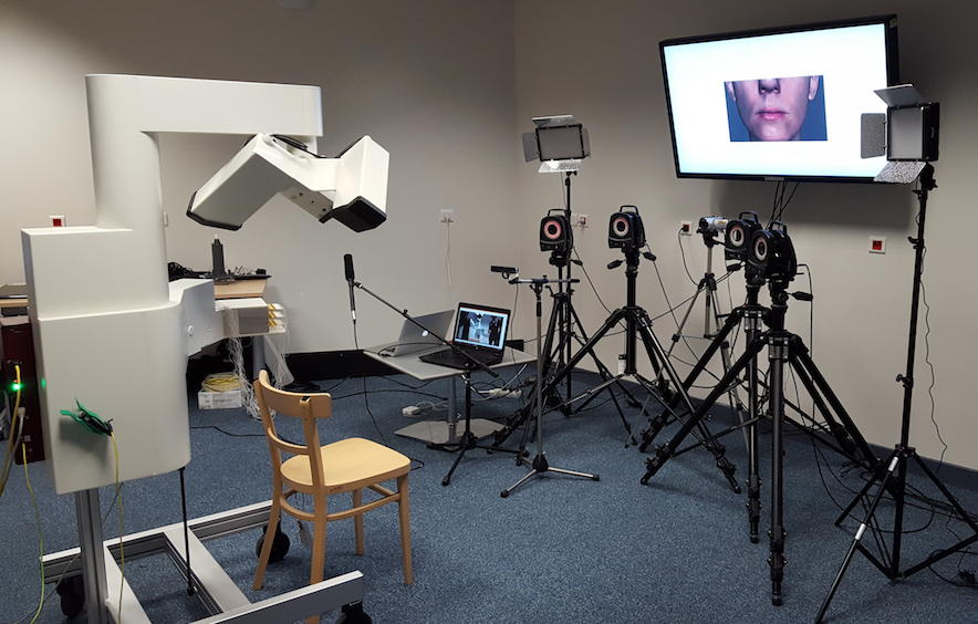 The acquisition room: the AG501 articulograph on the left, four Vicon cameras on tripods, lighting panels, a laptop, and a large screen showing a close-up of a speaker's mouth.