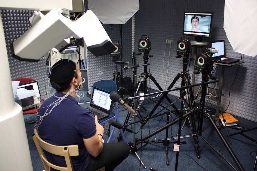 A speaker seated in the booth wearing the articulograph cap, sensor wires running from the face, facing the camera rig and a microphone during a recording session.