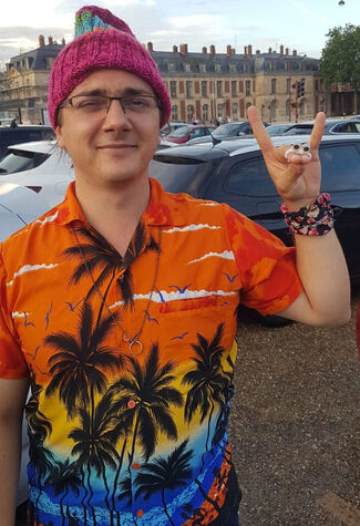 Photo of Guilhem wearing a hawaiian shirt and a colorful woolen cap. He has a pair of plastic eyeballs strapped around the middle and ring fingers of his left hand, turning said hand into some sort of puppet.